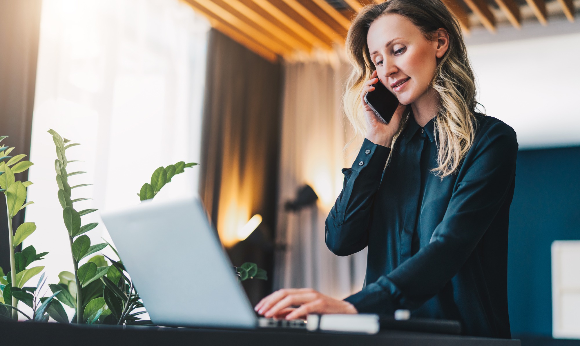 A woman with long hair talks on her phone while typing on a laptop, surrounded by greenery in a bright, modern workspace.