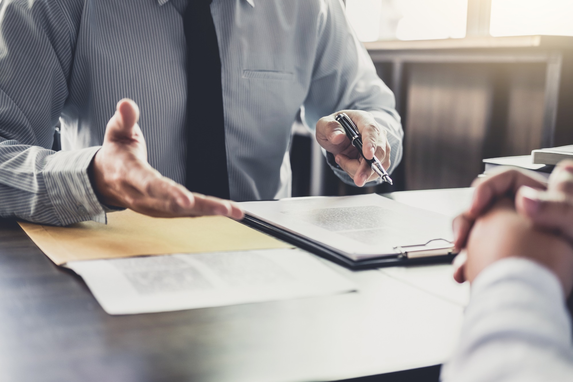 A business professional gestures while discussing documents with a client during a meeting, emphasizing important points with a pen.